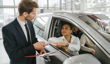 A car dealer hands keys to a woman sitting inside a vehicle in a car showroom.
