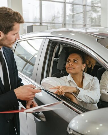 A car dealer hands keys to a woman sitting inside a vehicle in a car showroom.