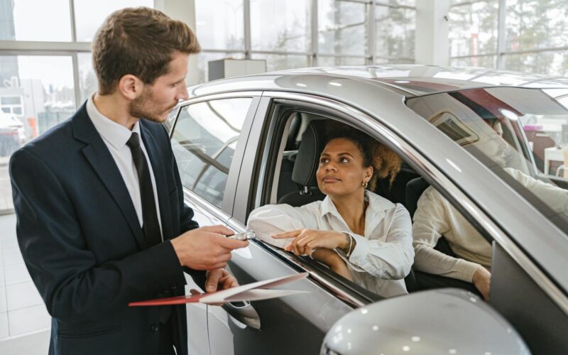 A car dealer hands keys to a woman sitting inside a vehicle in a car showroom.