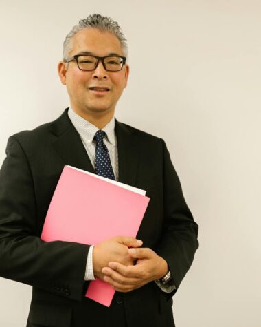Confident businessman in black suit holding pink folder, smiling at the camera indoors.