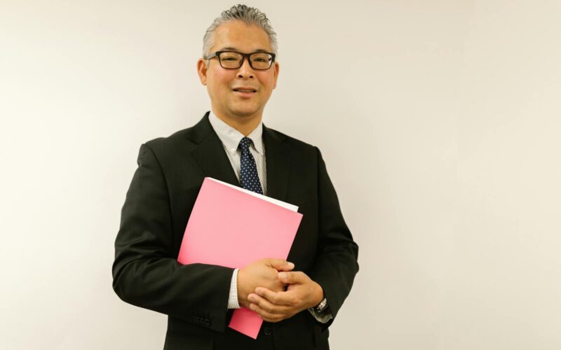 Confident businessman in black suit holding pink folder, smiling at the camera indoors.